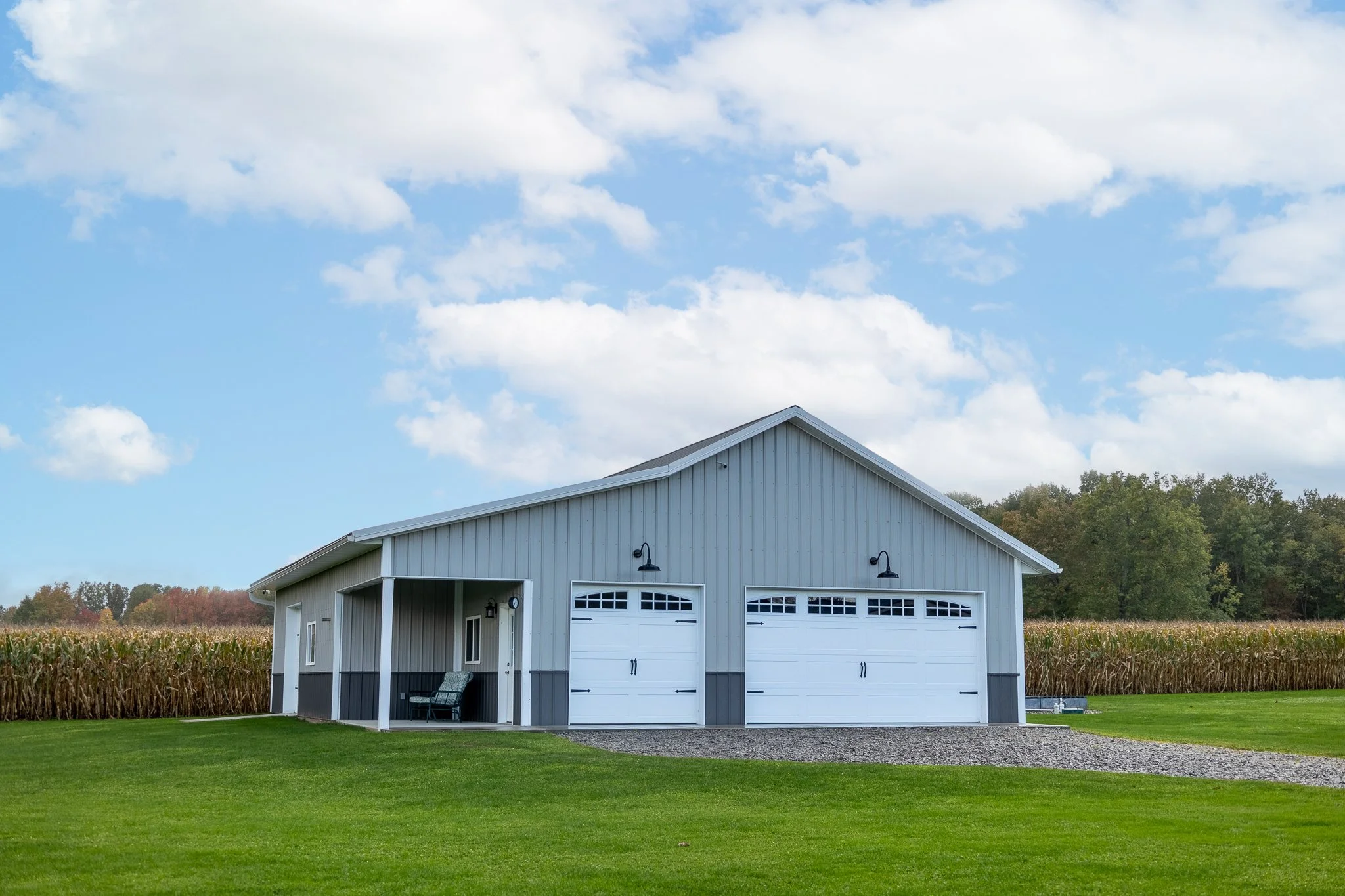 A grey barn with white garage doors, a small porch with a bench, surrounded by green grass and a cornfield, under a partly cloudy sky.