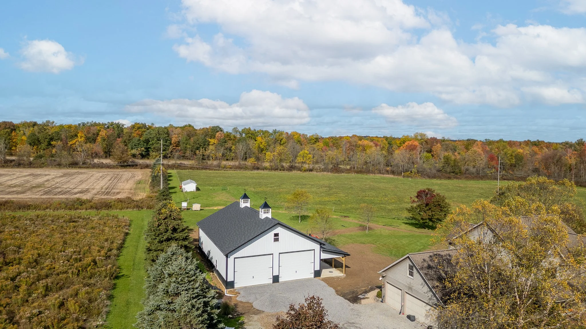 Aerial view of a farm with a white barn, a gray house, a field, and trees with fall foliage under a partly cloudy sky.