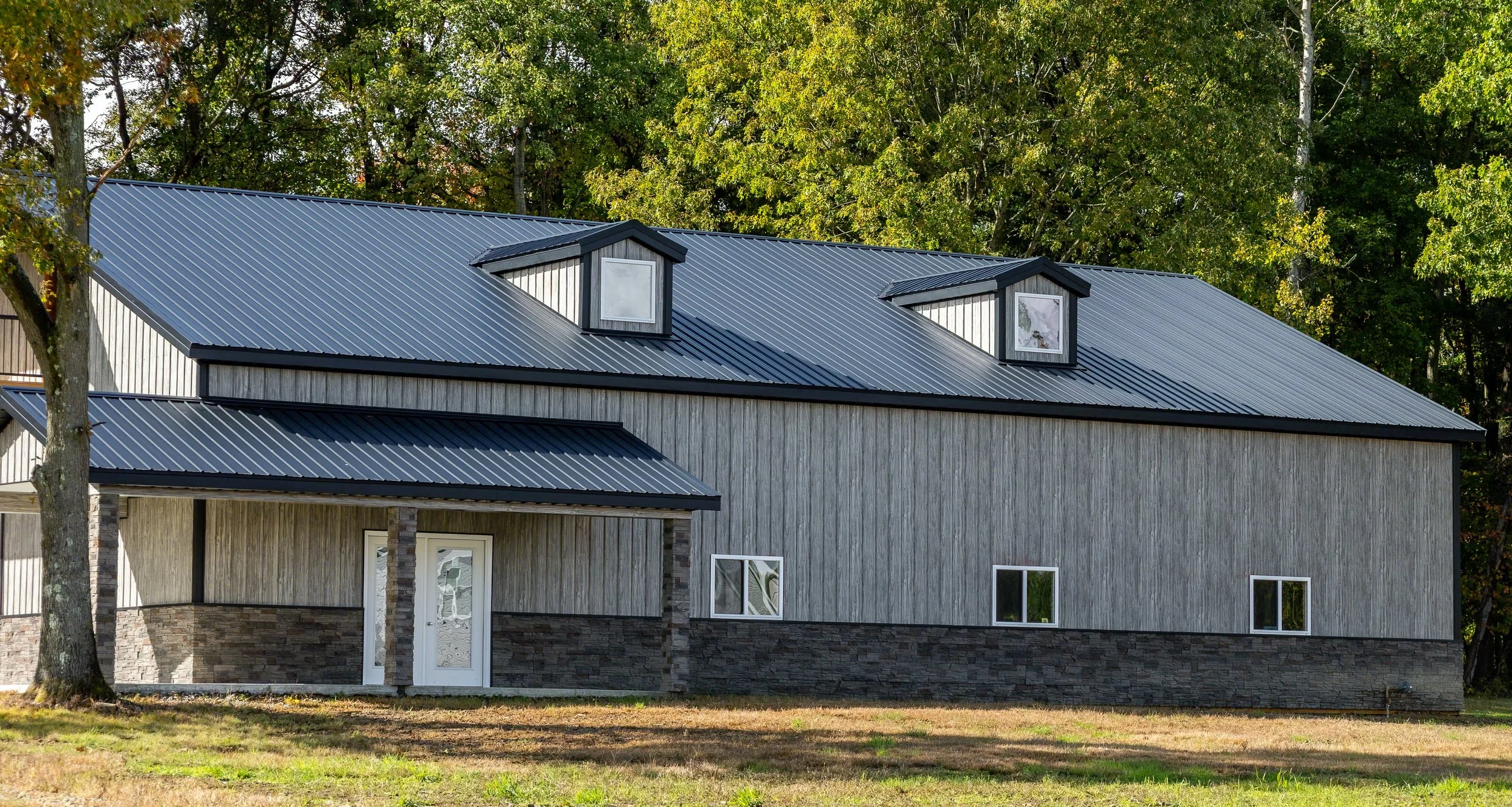 A two-story house with a metal roof, vertical wooden metal siding, stone metal siding accents at the bottom, and small windows, surrounded by trees.