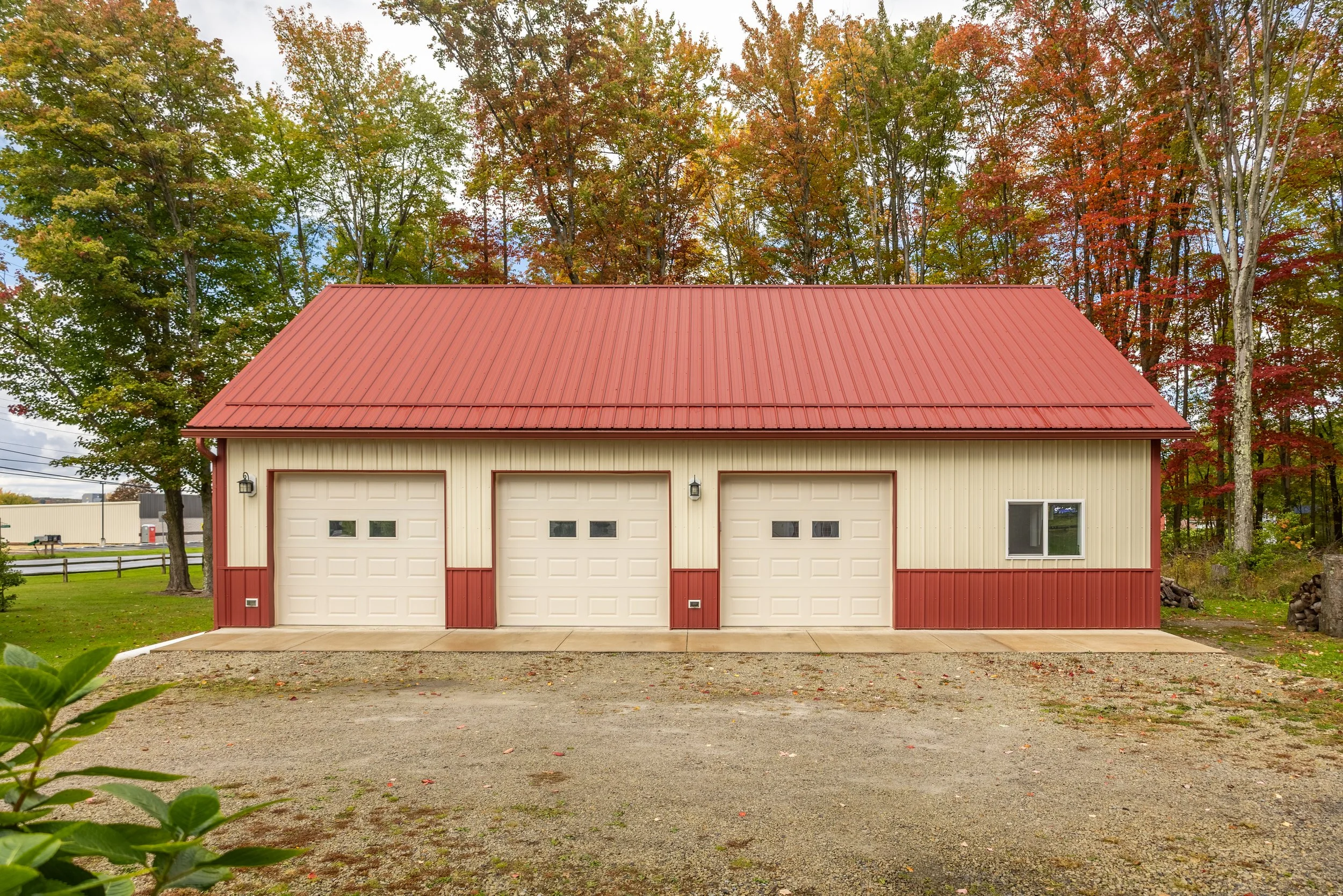 A large beige and red metal garage with three white doors, two small windows near the doors, and surrounded by trees with fall foliage.