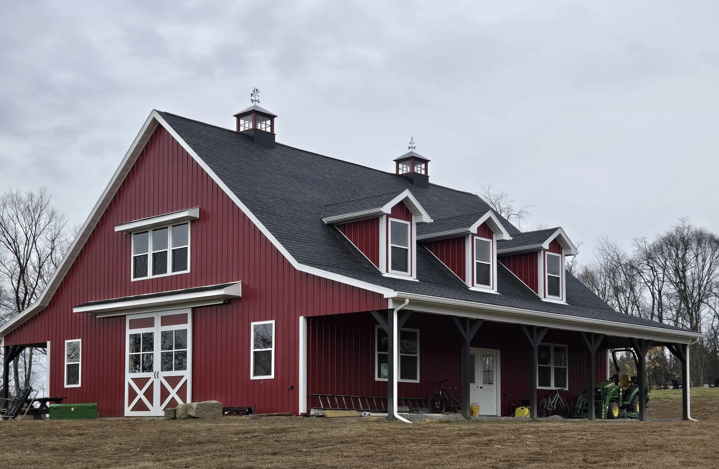 A large red barn with white trim, black roof, and multiple windows, situated in a rural area with leafless trees in the background, and various equipment and bicycles outside.