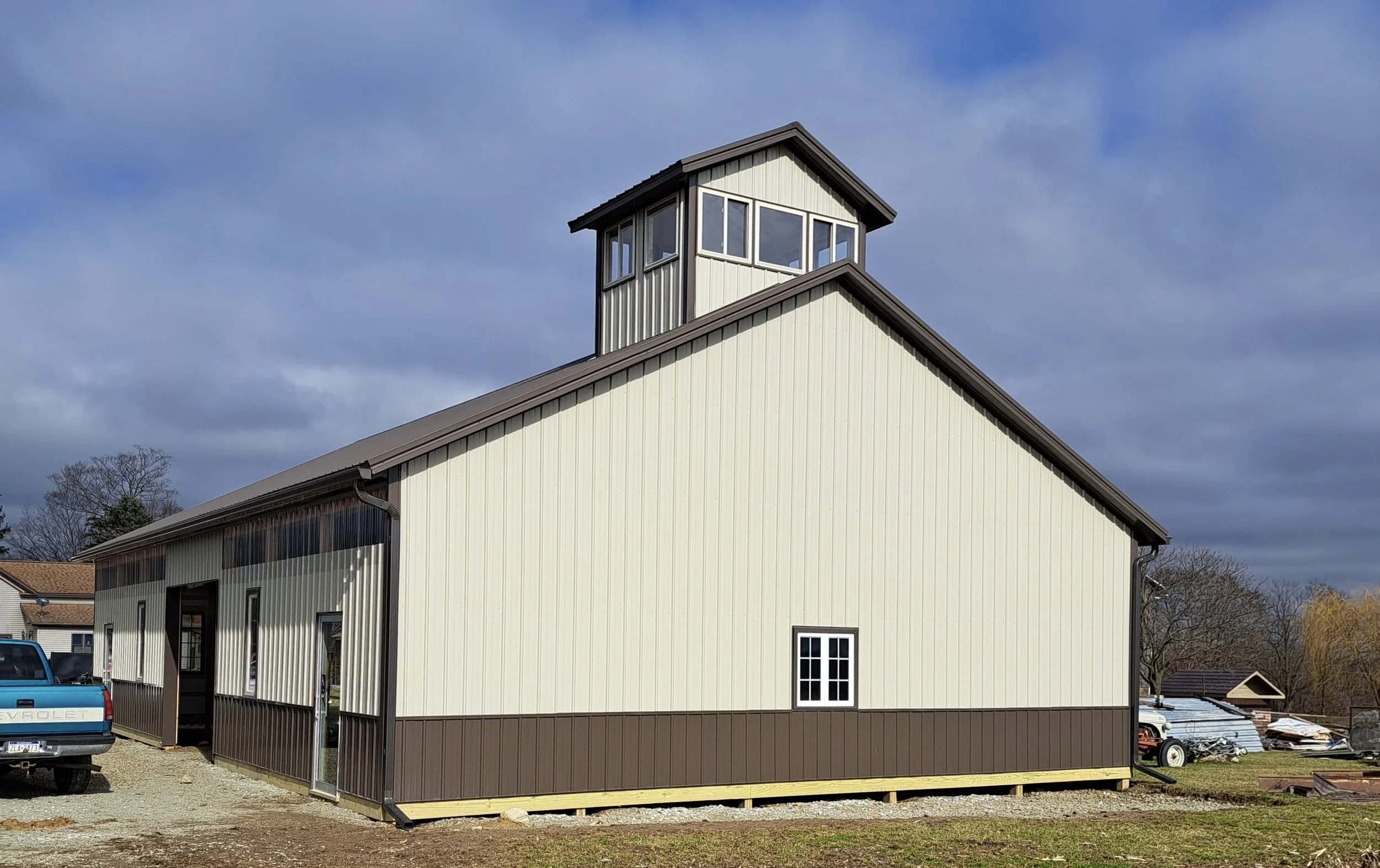 A large beige and brown building with a small tower on top, set against a cloudy sky, with a blue truck in the foreground and trees and other structures in the background.