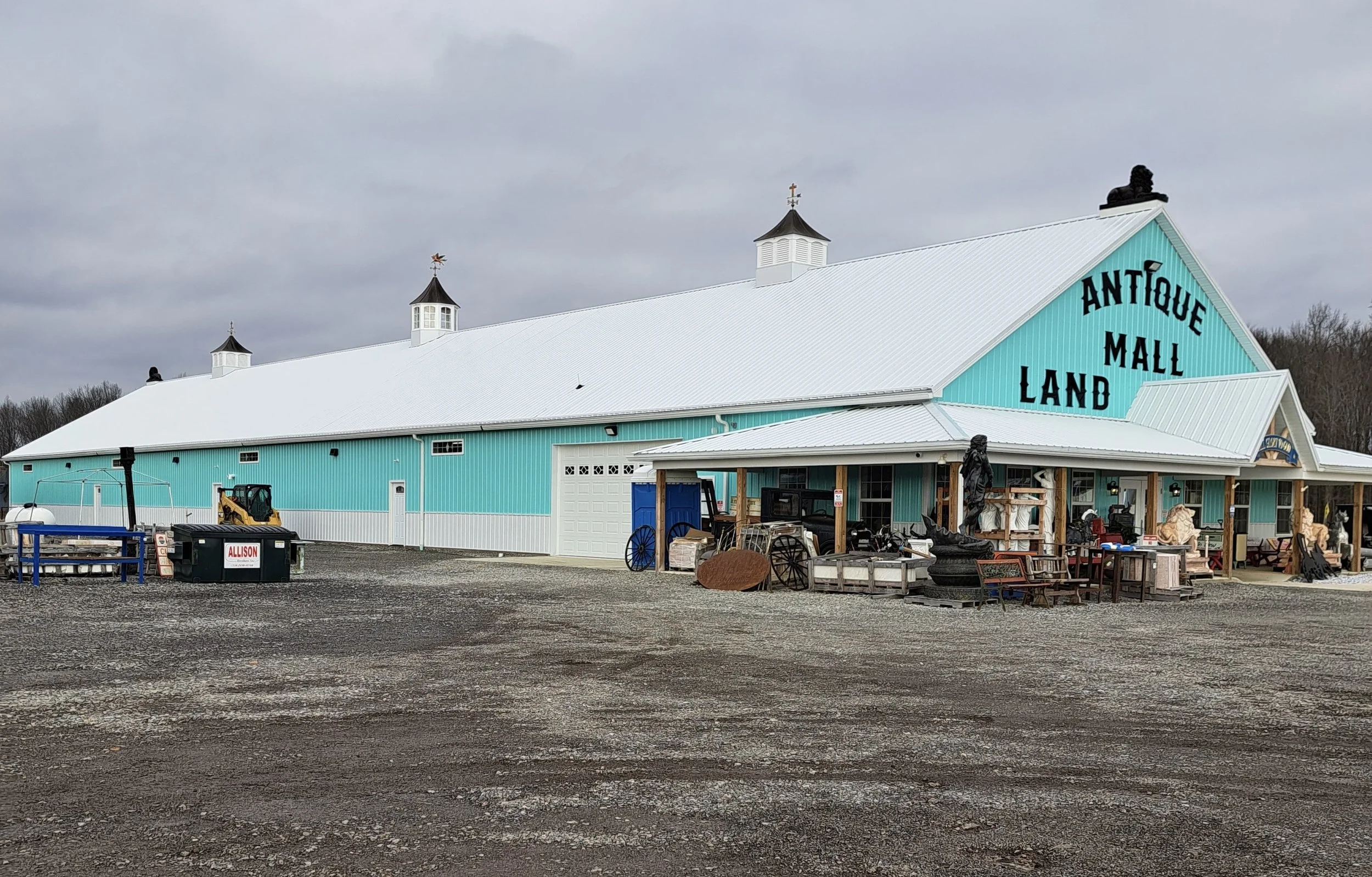 Large teal-colored barn with the words 'Antique Mall Land' written on the side, surrounded by various antique items and outdoor furniture, under an overcast sky.