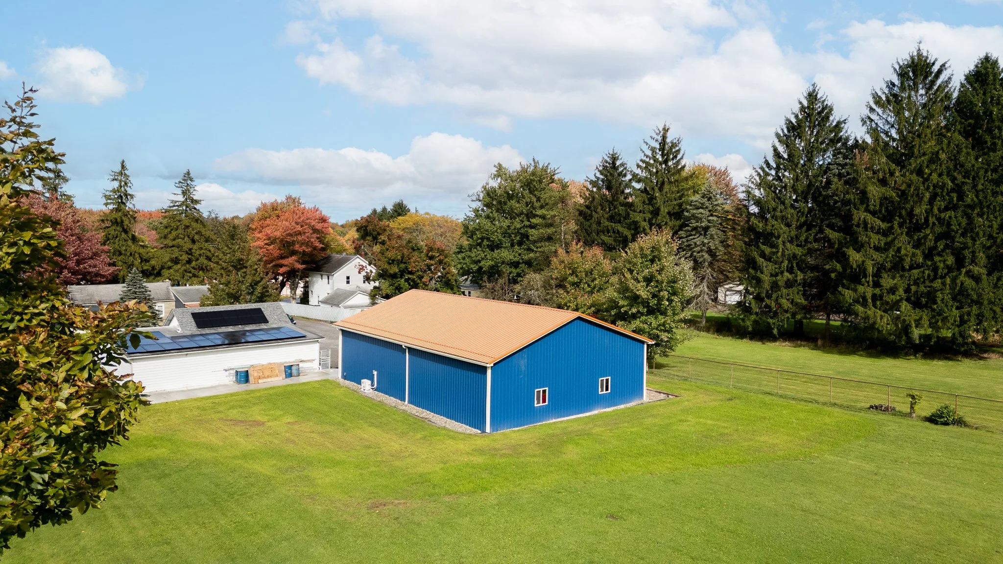 A blue metal building with a tan roof in a grassy yard surrounded by trees and neighboring houses.