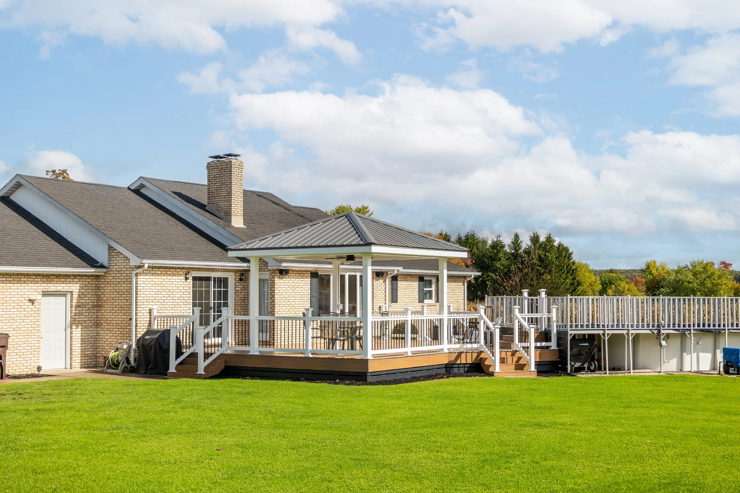 Back of a brick house with a gray metal roof, a brick chimney, and a large wooden deck with a covered gazebo, railings, stairs, and outdoor furniture, surrounded by a green lawn and trees under a partly cloudy sky.