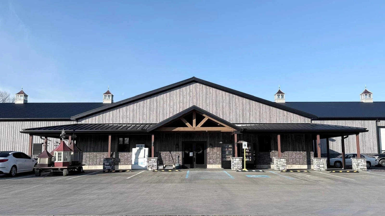Exterior view of Rolling Ridge Metals LLC with a large parking lot, metal siding, a gable roof, and cupolas on top of the roof, under a clear blue sky.