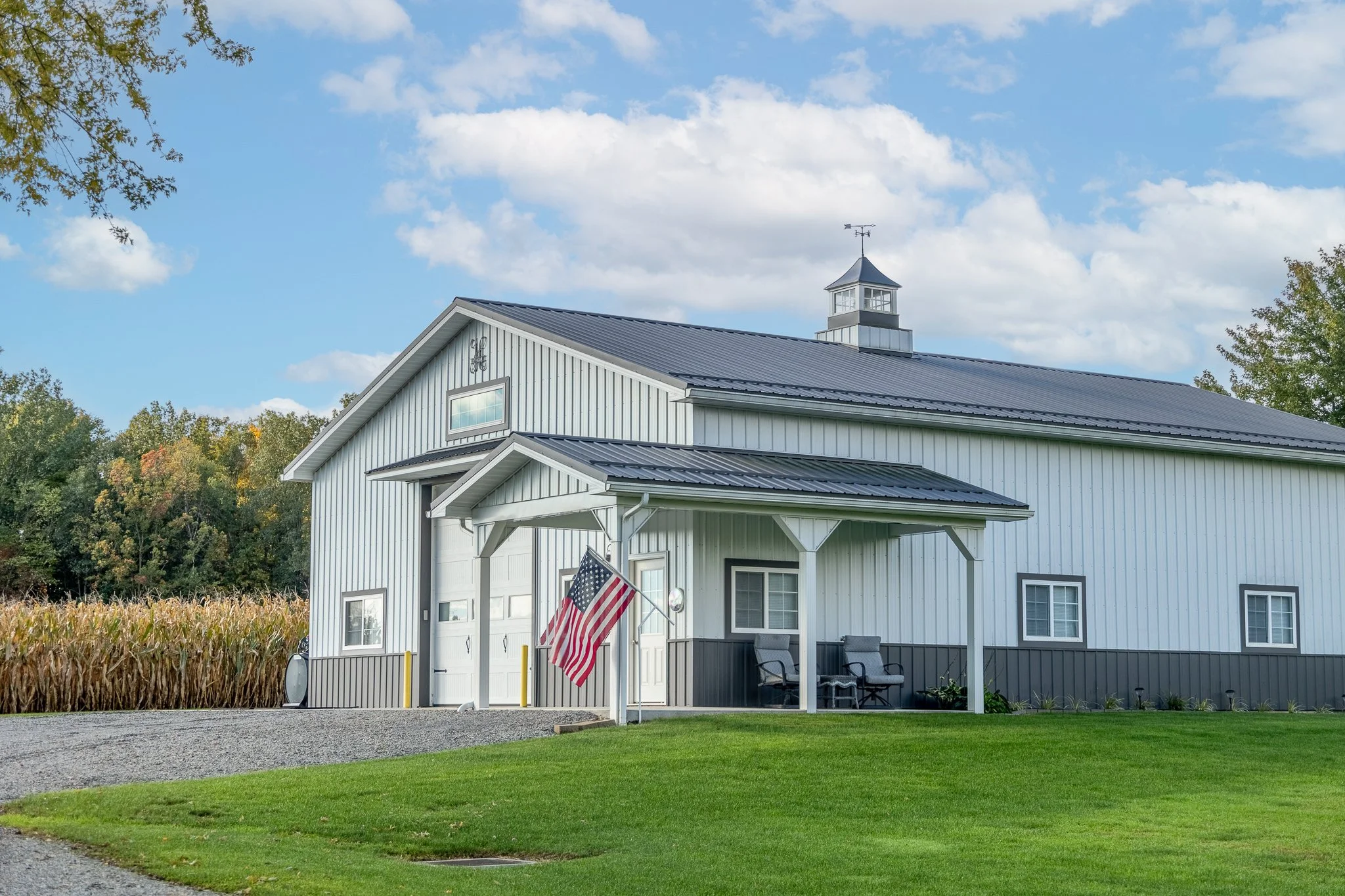 A white barn with a dark roof, American flag, gravel driveway, and green lawn surrounded by trees and cornfield, under a partly cloudy sky.