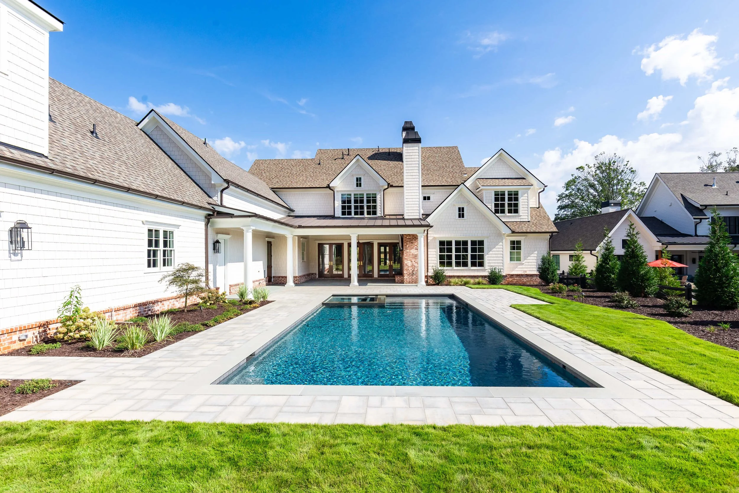 Backyard of a large white house with a swimming pool, green lawn, and landscaped garden with small trees and shrubs under a blue sky
