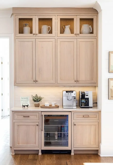 Kitchen cabinet with upper glass doors displaying white teapots and pitchers, lower cabinets, a countertop with a coffee machine and mini fridge, and framed artwork on the wall.