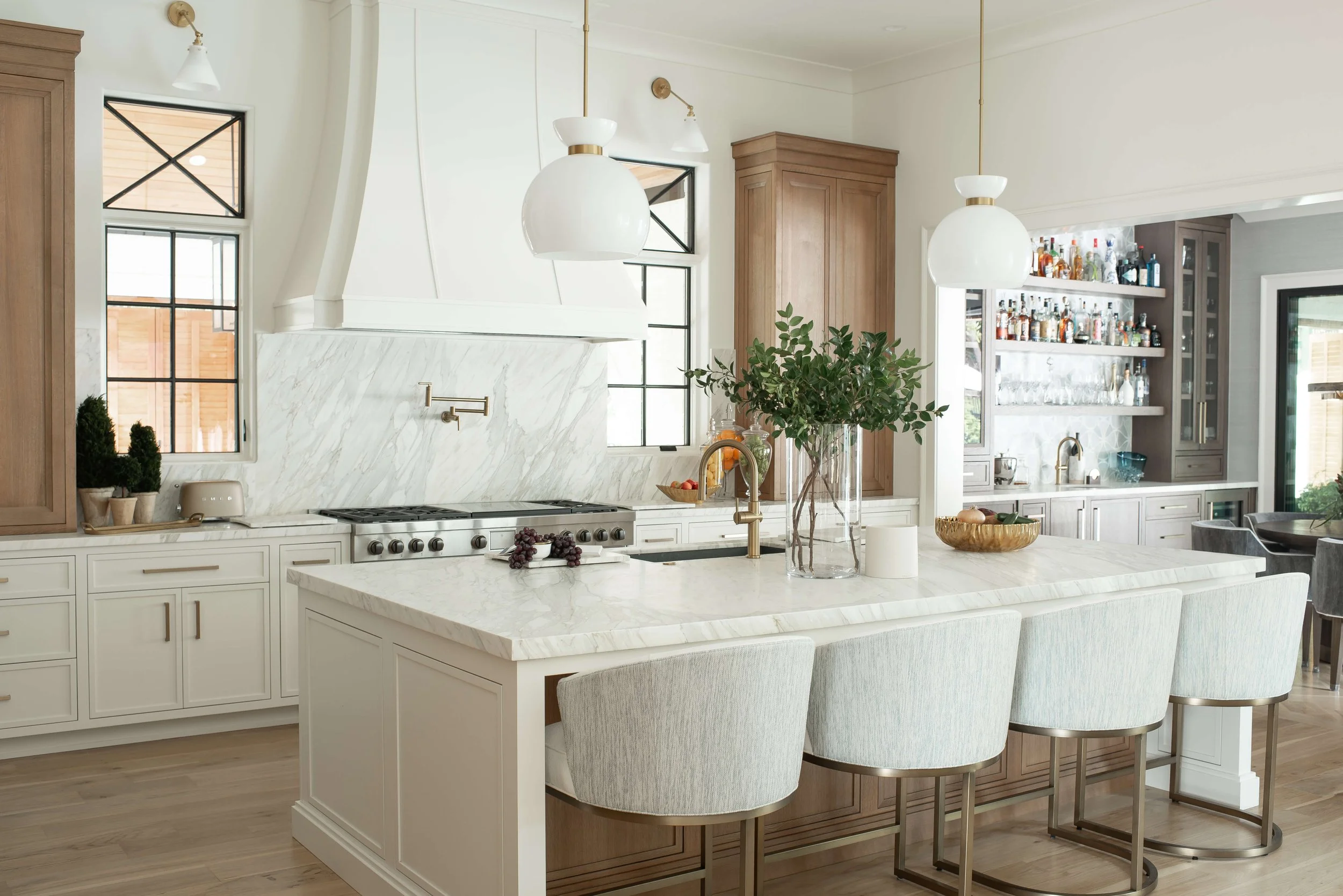 Modern kitchen with white marble island, wooden cabinets, and black-framed windows. There are pendant lights above the island, a bar area with bottles, and decorative plants.