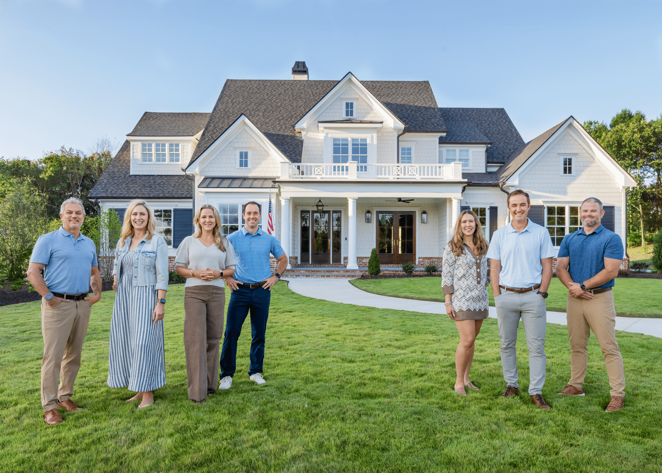 The Lehigh Homes team stands in front of a large white house with a manicured lawn and trees in the background.