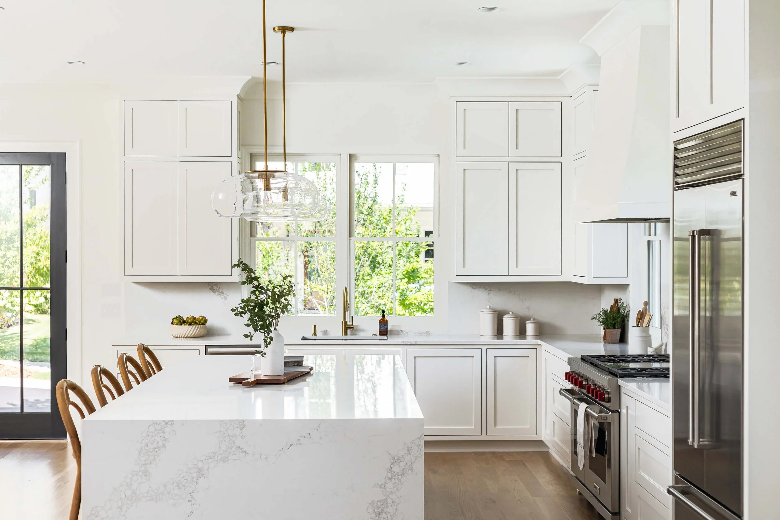 Bright white modern kitchen with island, wooden chairs, stainless steel appliances, and large windows showing greenery outside.