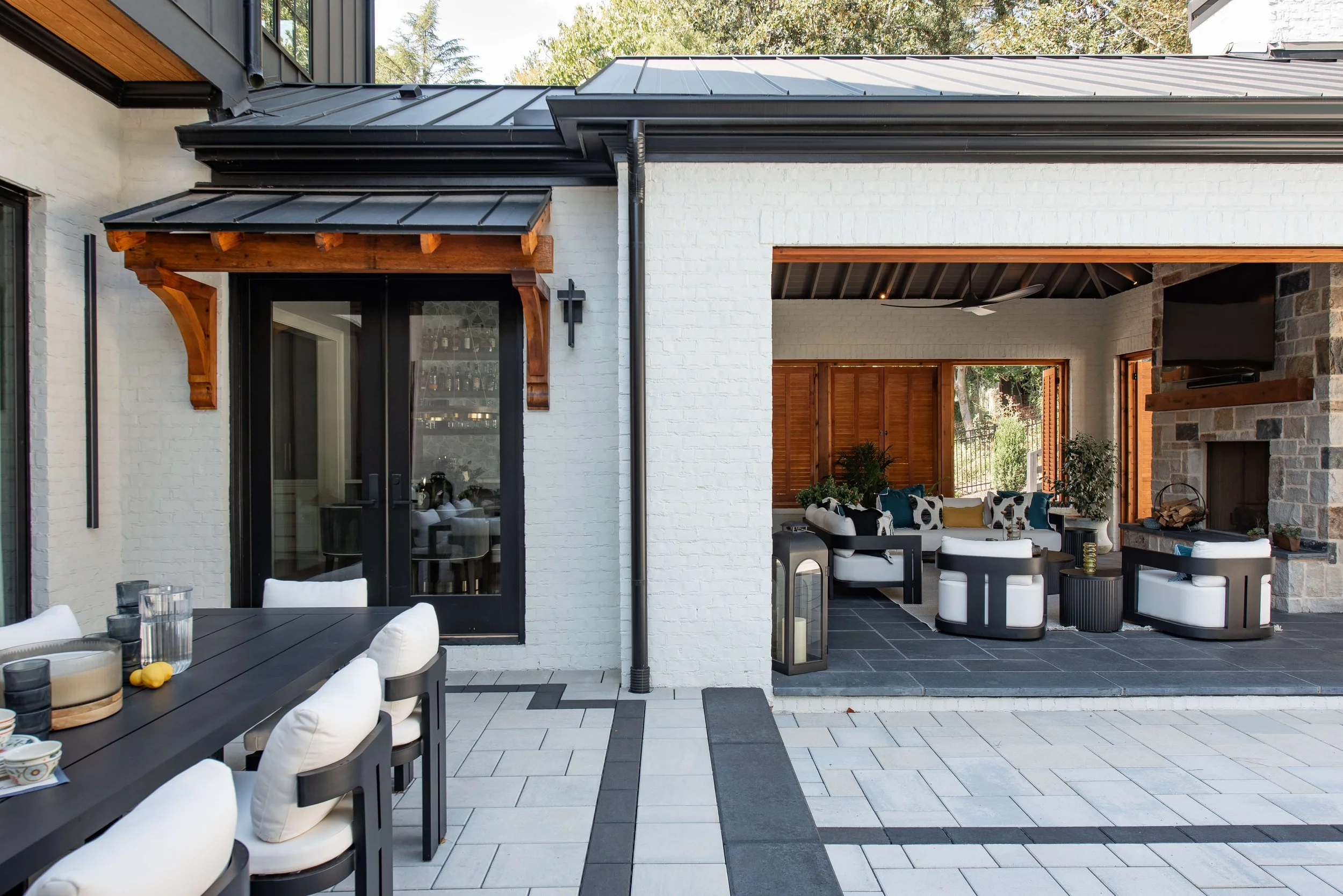 Outdoor patio with dining area on the left featuring a black table and white cushioned chairs, and a lounge area on the right with white and black furniture, a fireplace, and plants under a metal roof, with a white brick house in the background.