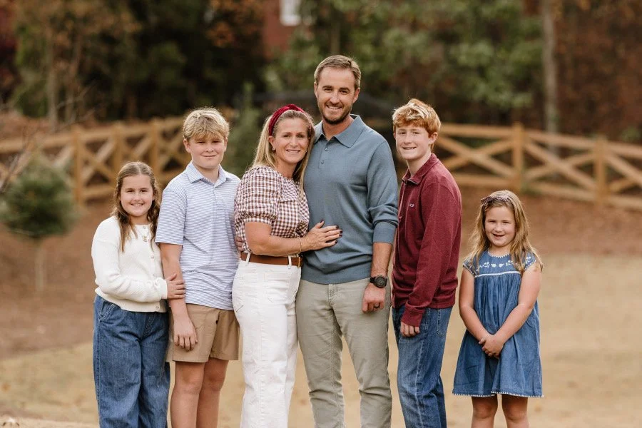 Brendan Walsh and his family of 6 stand smiling in front of a wooden fence in the background.