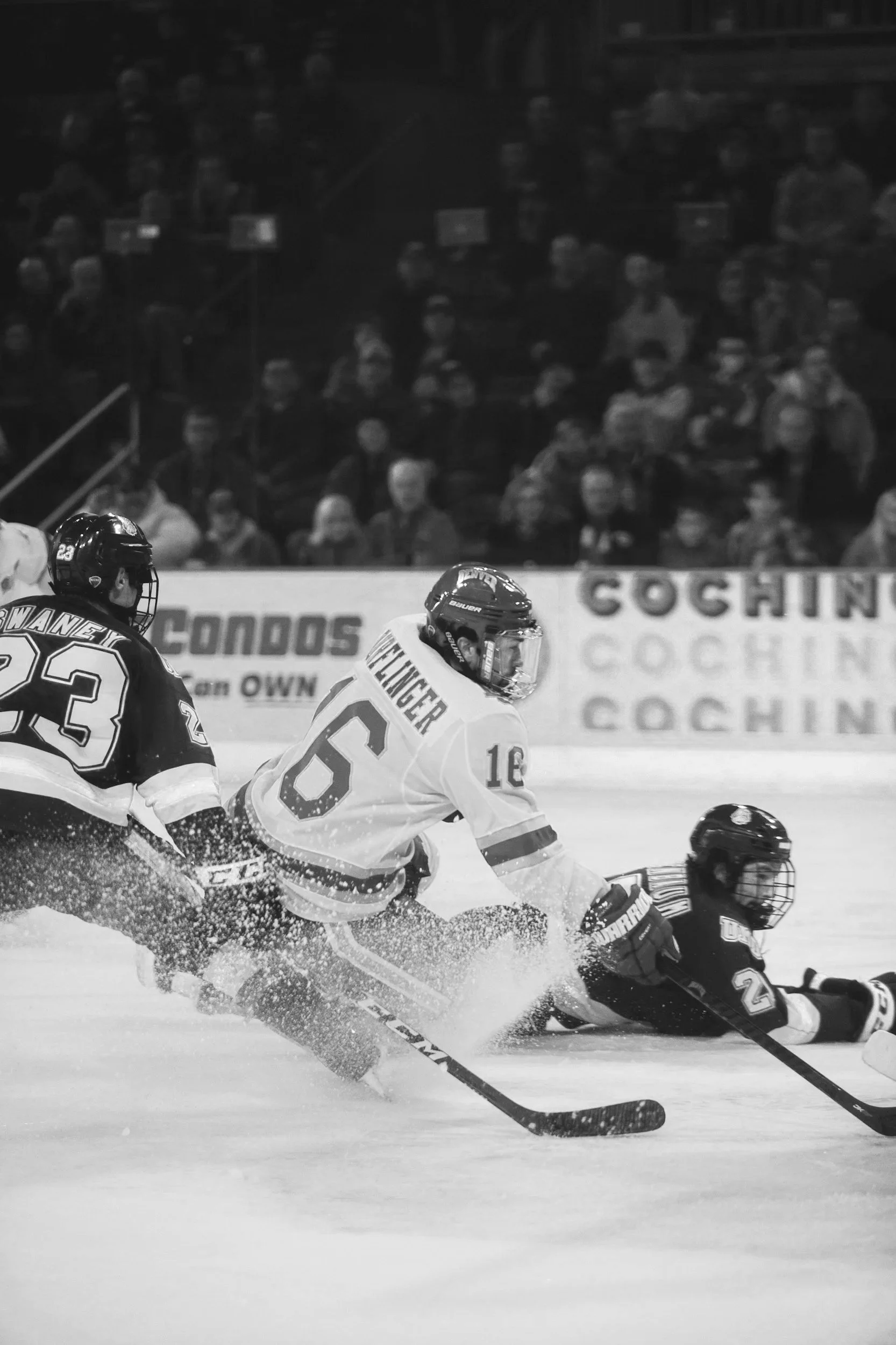 Black and white photo of an ice hockey game showing two players and a goalie fighting for the puck, with spectators in the background.