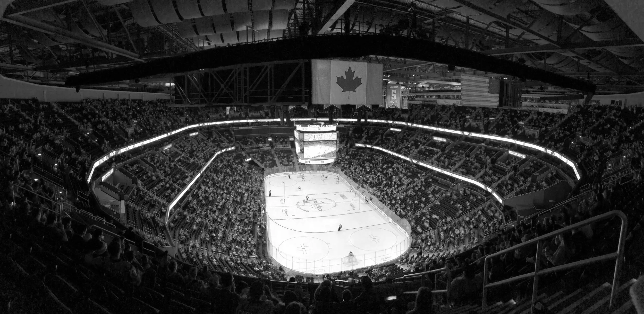 Black and white photo of an indoor ice hockey arena filled with spectators, with a view from the stands and players on the ice.