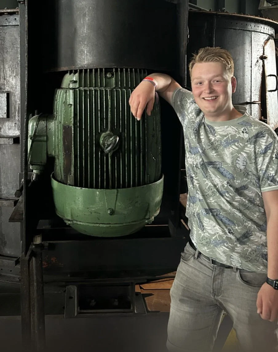 Een jonge man lacht en poseert met zijn arm om een ​​grote groene, vintage machine of motor, in een industriële omgeving of werkplaats.