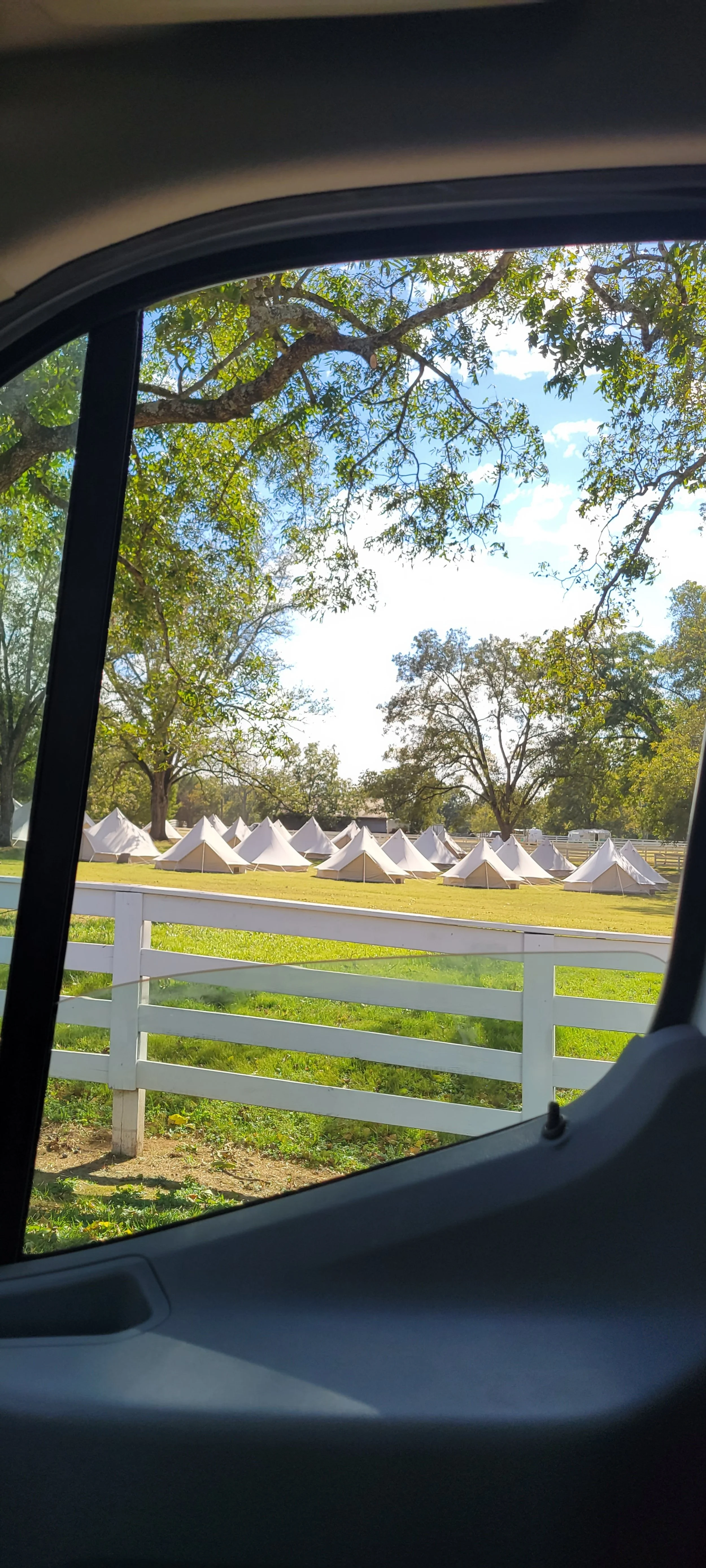 A scene viewed from inside a vehicle showing a grassy field with white tents set up in rows, several trees, a white fence, and a partly cloudy sky.