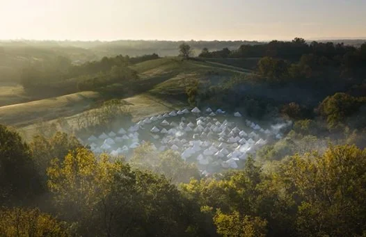 A campsite with numerous white tents arranged in a circular pattern among green trees in a hilly landscape, with morning sunlight and light mist.