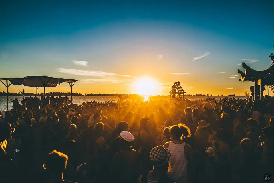 Crowd of people gathered outdoors by a river during sunset with umbrellas and structures