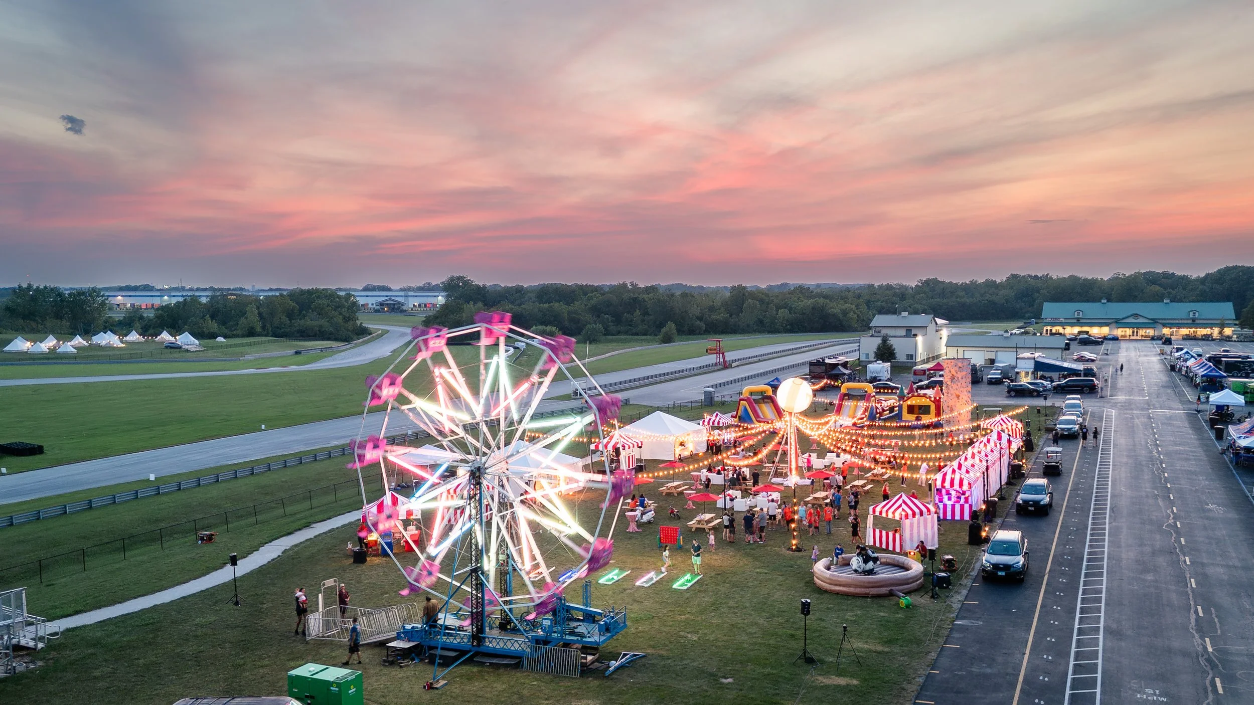 An amusement park with a Ferris wheel, carousel, and tents, set up in a large open field at dusk, with lights illuminating the rides and attractions.