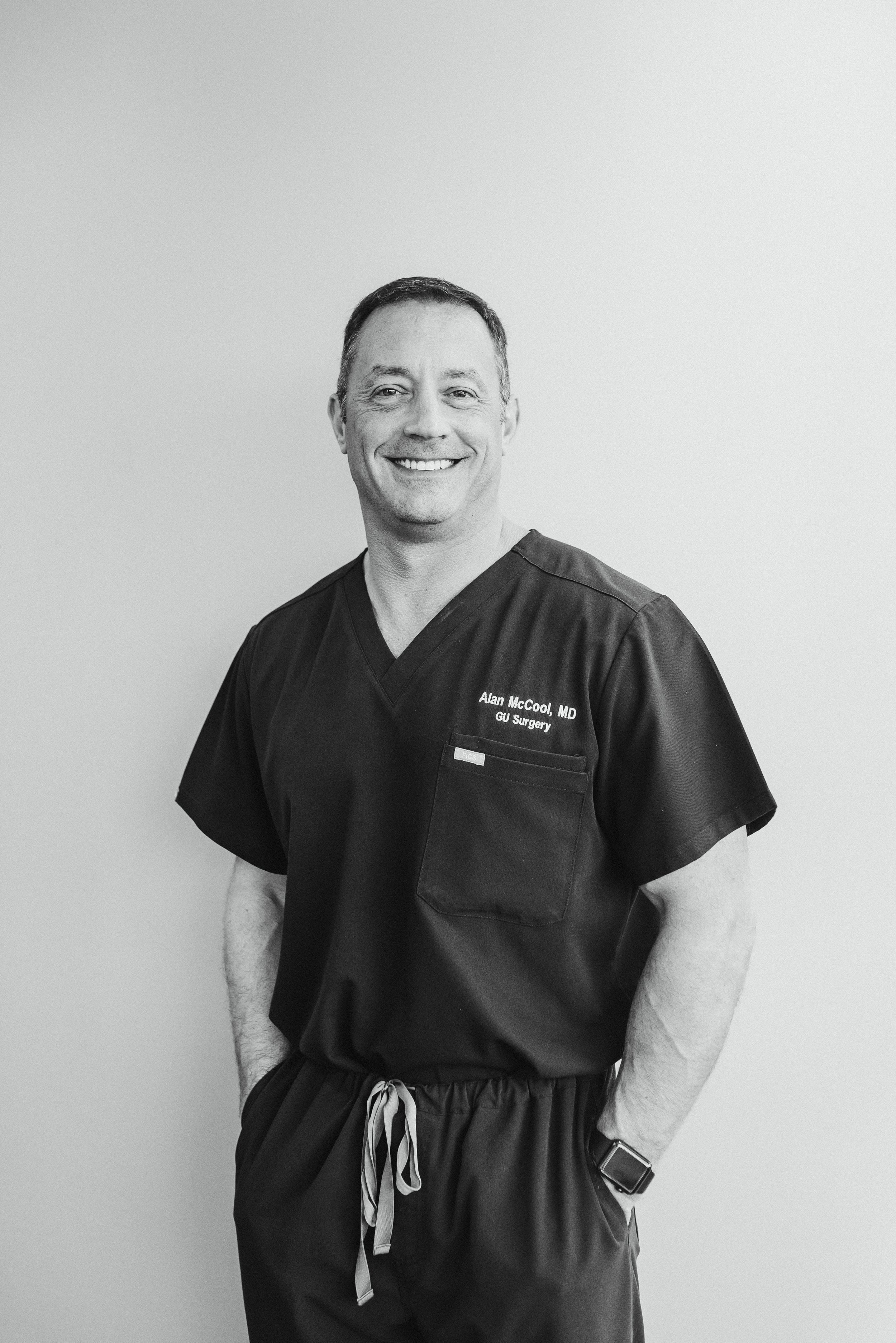 A smiling male doctor in medical scrubs with a name tag, standing against a plain wall, with his hands in his pockets.