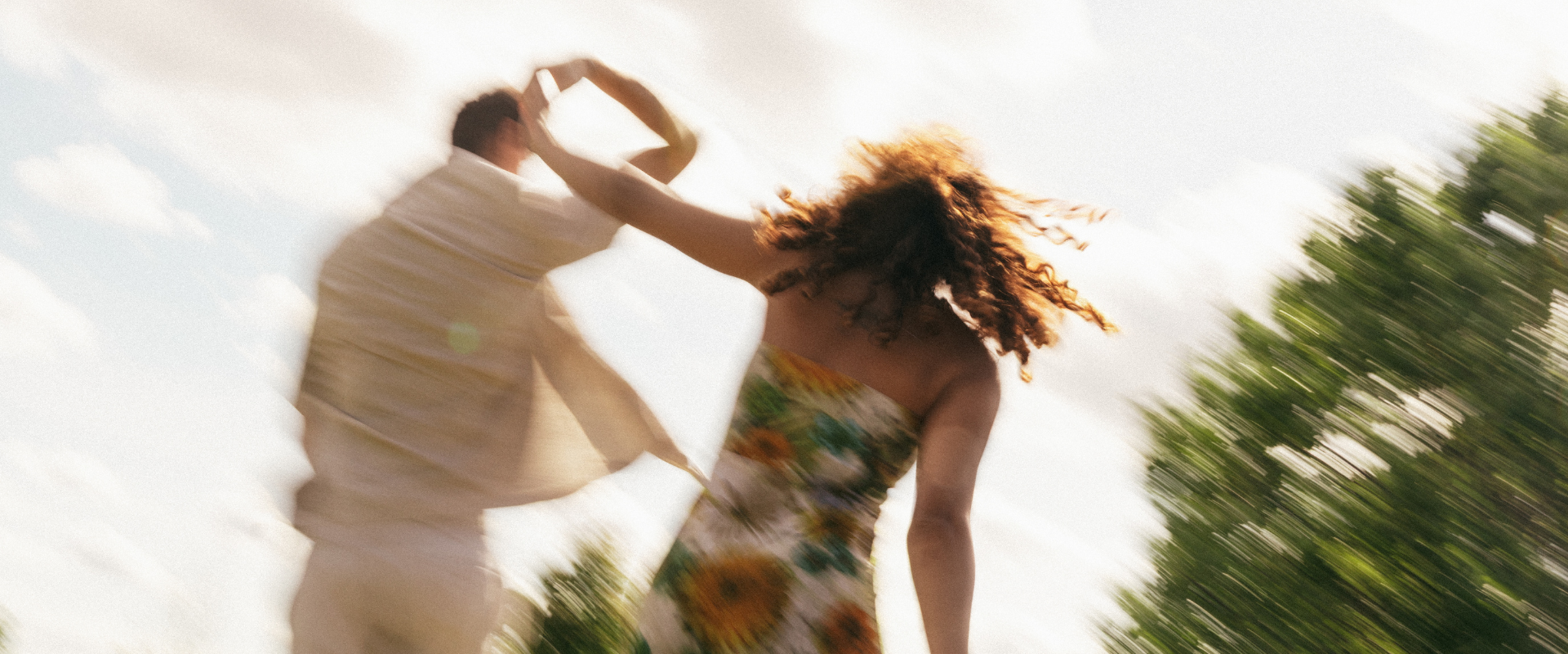 A couple dancing outdoors under a bright sky with blurred trees and clouds, with motion blur indicating movement.