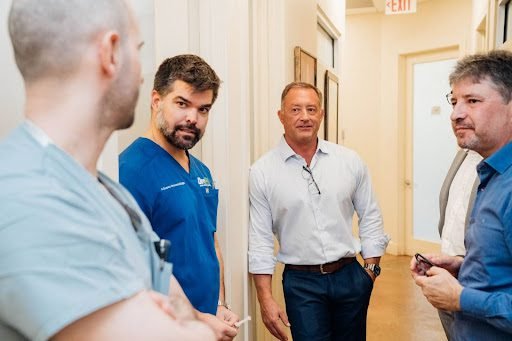 Four men engaged in conversation in a clinic hallway, three facing the camera and one with his back turned, dressed in professional attire.