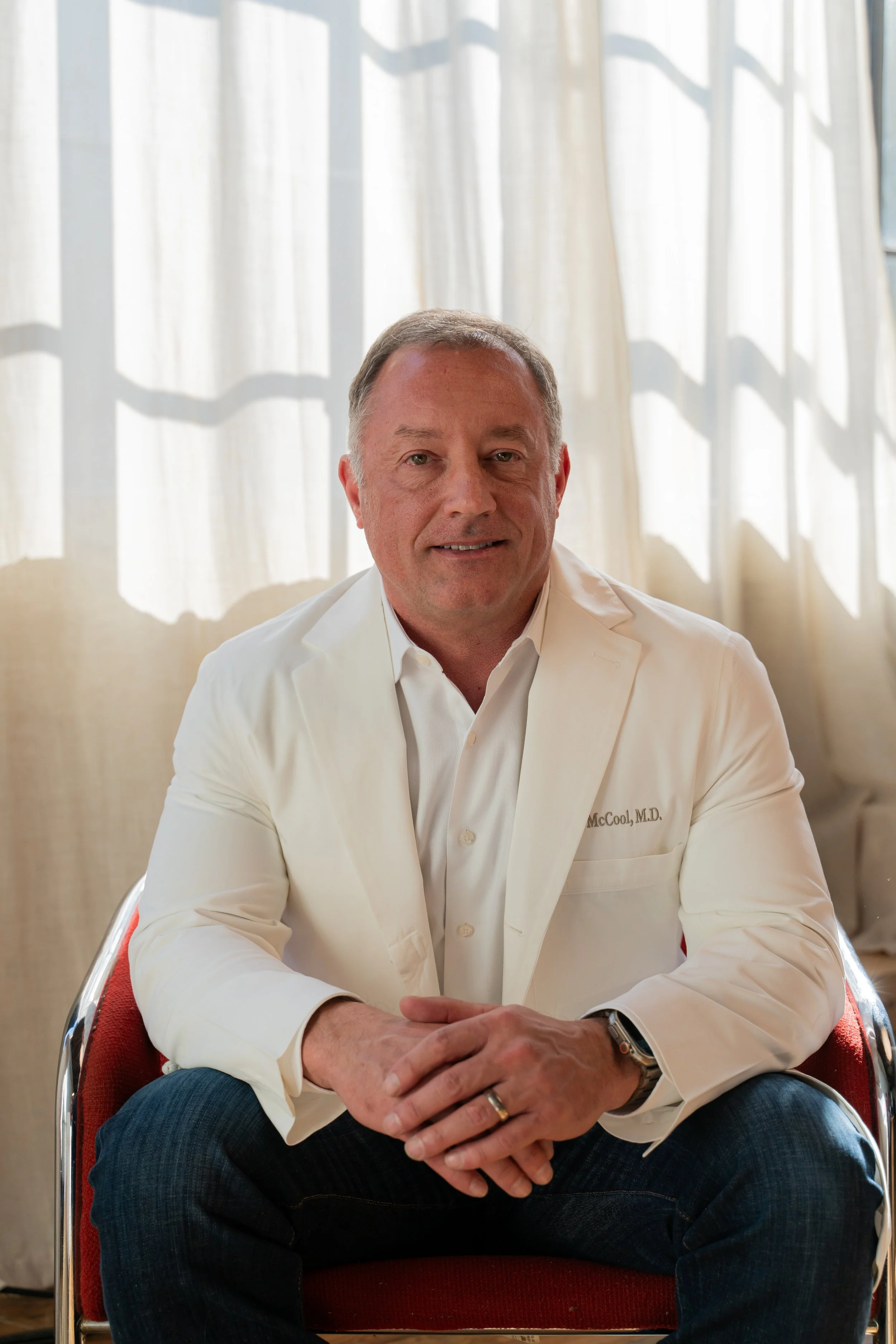 A middle-aged man with light skin, short gray hair, dressed in a white doctor’s coat with a name tag, sitting on a red chair in front of a window with sheer curtains.