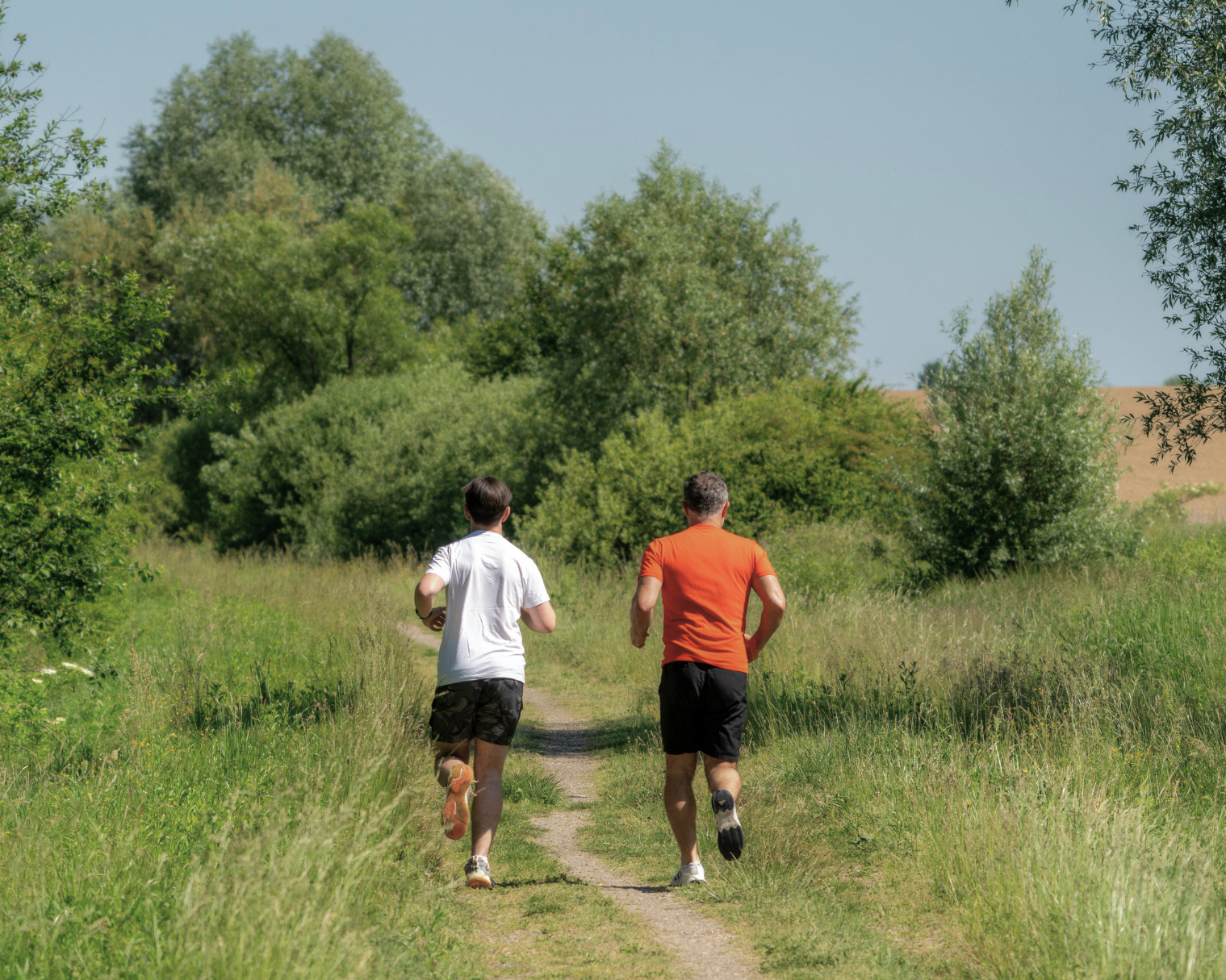 Two men jogging on a dirt path through a green field near trees under a clear blue sky.