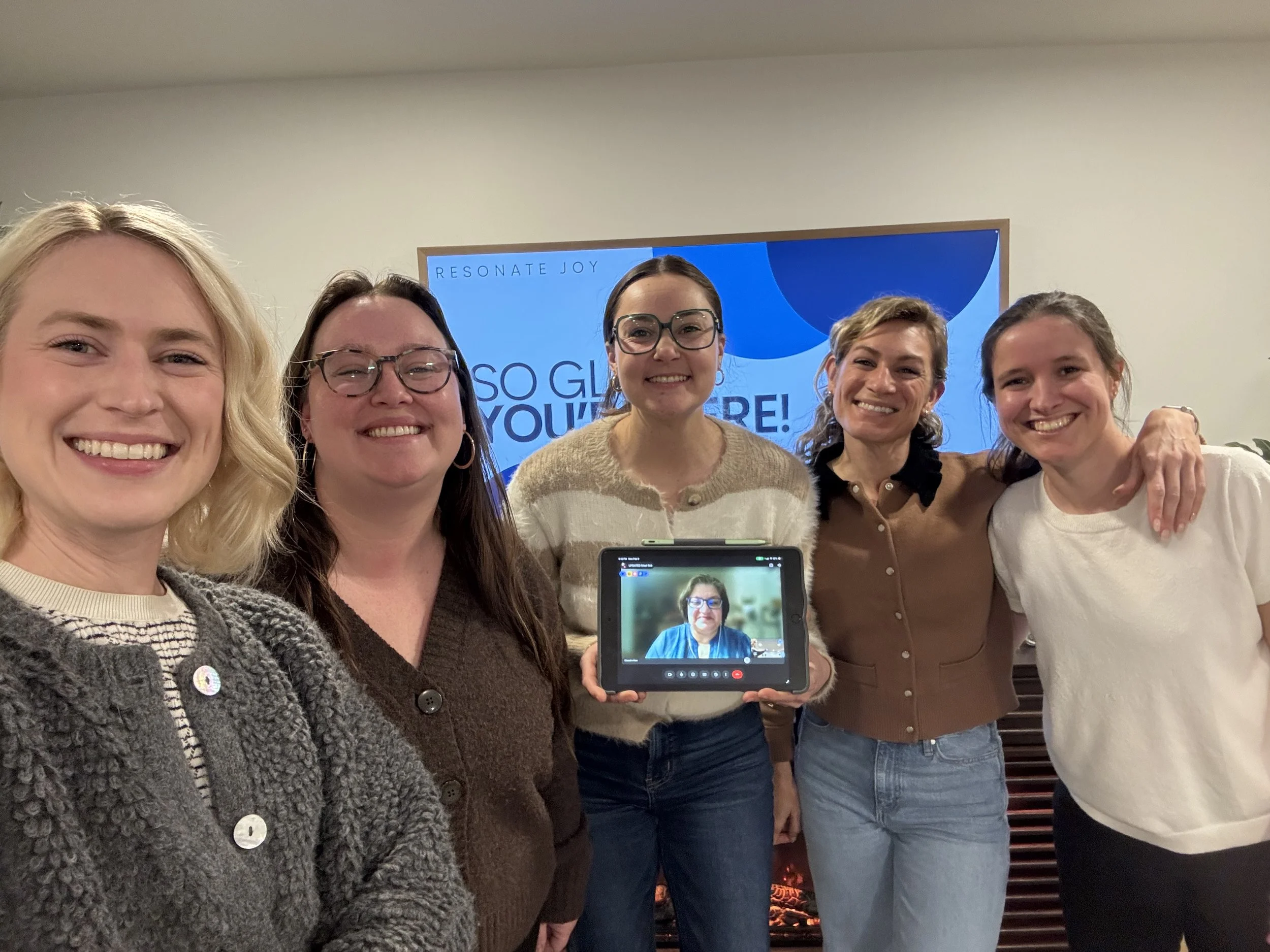 Resonate Joy's leadership team at a board meeting: five women are standing closely together, smiling at the camera. One woman in the center is holding a tablet with a person on the screen in a video call.