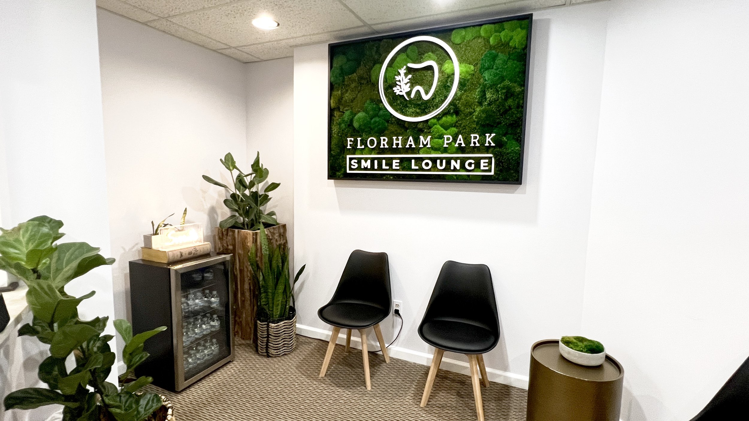Interior of Florida Park Smile Lounge with two black chairs, potted plants, a small cabinet with glasses, and a large screen with the lounge's logo.