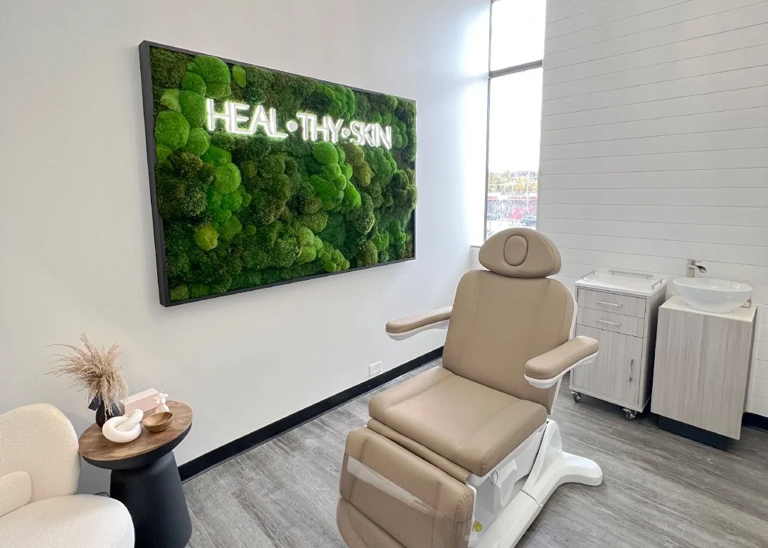 A treatment room with a beige medical chair, a small side table with decorative items, a wall-mounted digital display with the words "HEAL·THY·SKIN" in neon lights, a white cabinet with a wash basin, and a large window allowing natural light.