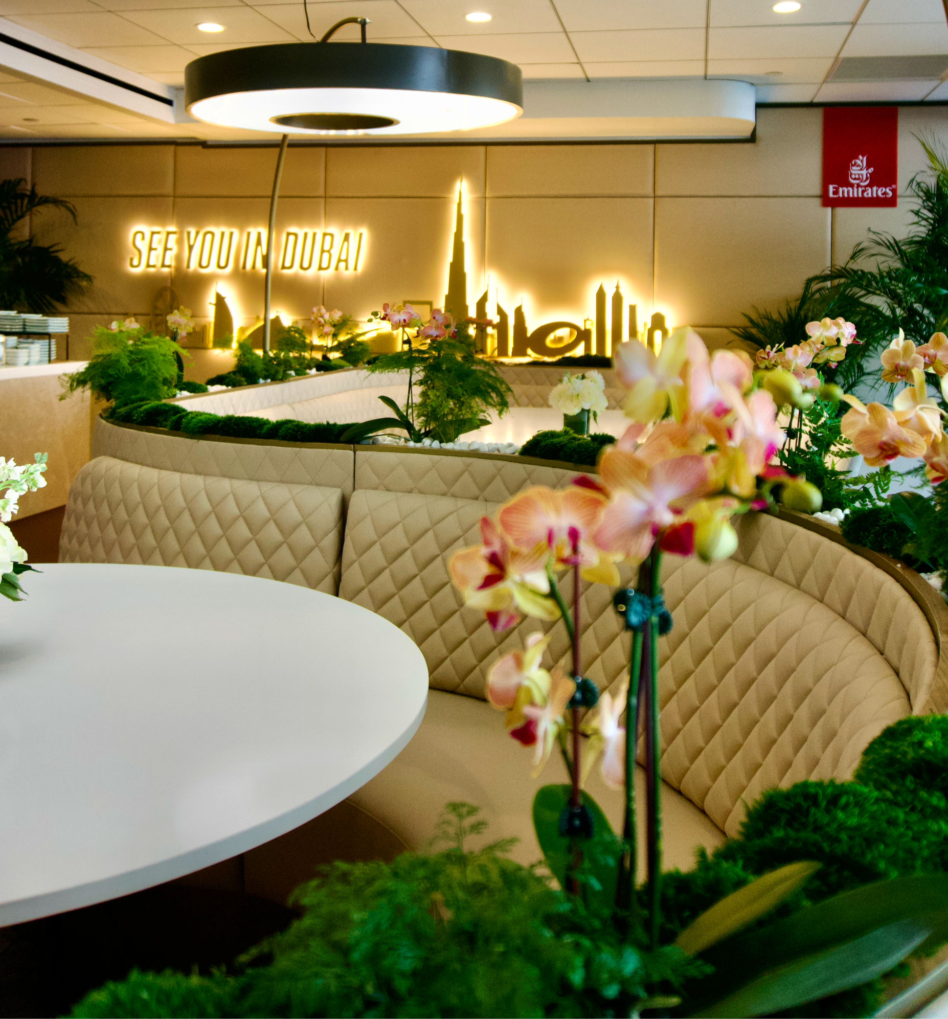 Indoor seating area with beige quilted booth, white table, pink orchids, green plants, illuminated sign reading "See you in Dubai", and Emirates logo in background.