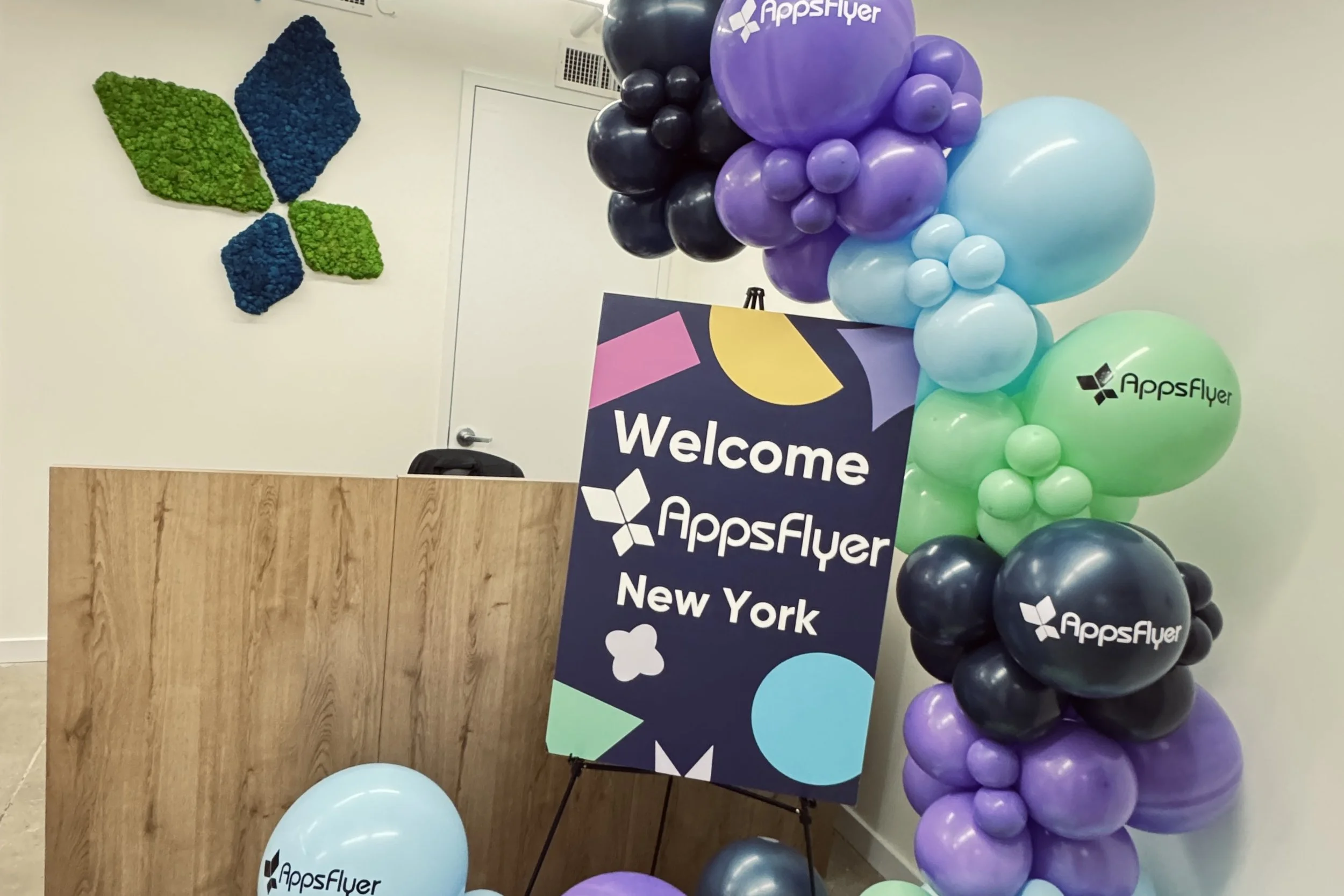 A reception area with a wooden desk, a black chair, a decorative wall art made of green and blue moss, a colorful 'Welcome' sign for AppsFlyer in New York, and a large arrangement of balloons in purple, black, light blue, green, and dark blue with th
