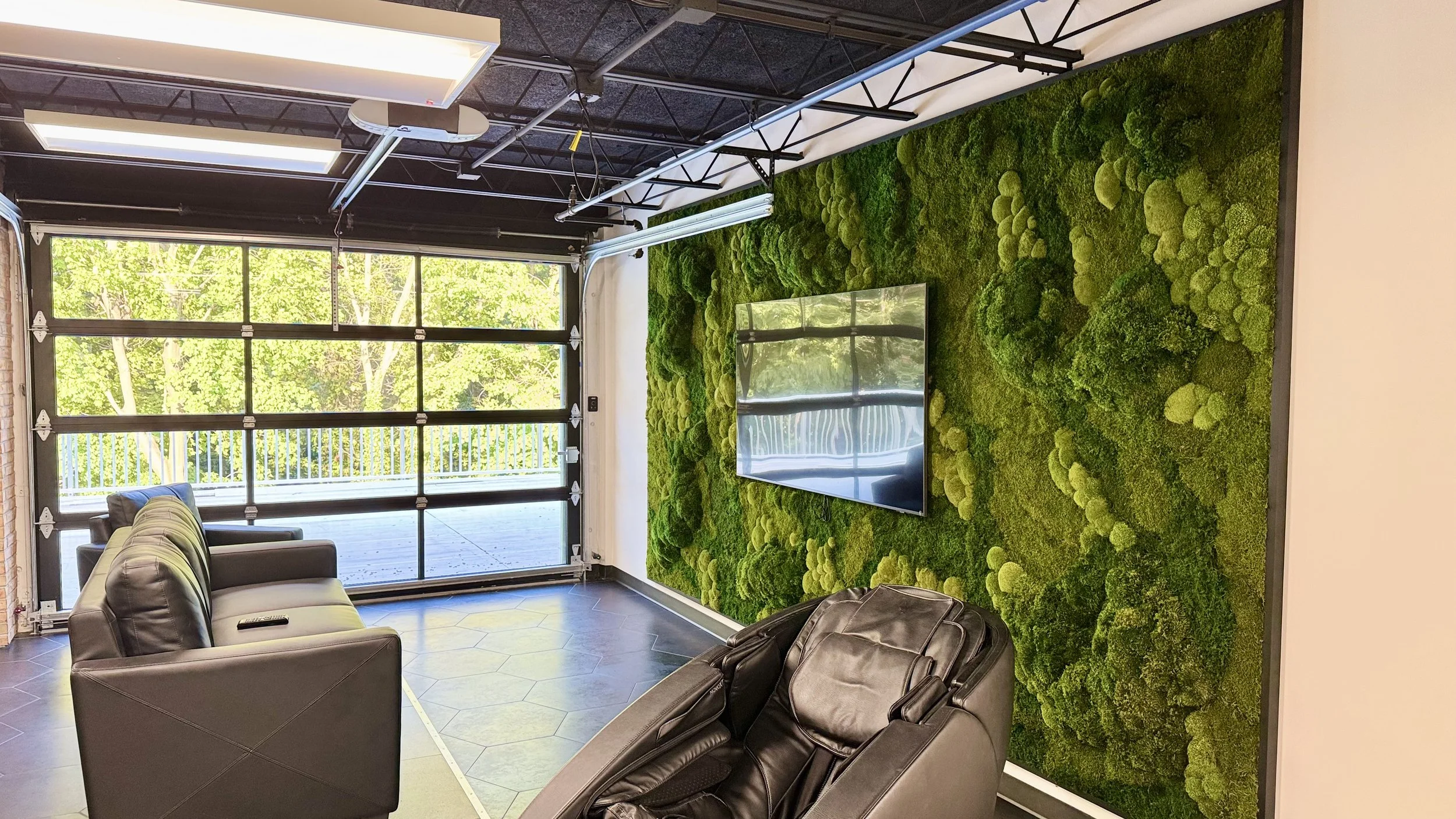 Living room with black leather recliner, a gray leather couch, and a wall covered in green moss with a mounted flat-screen TV. Large windows with a garage door overlooking green trees outside.