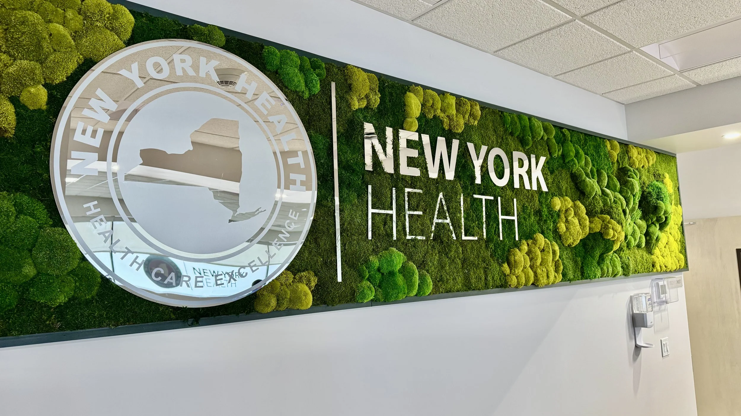 Interior wall display with green moss and plants, featuring the logo of New York Health and the text 'NEW YORK HEALTH' in white letters.