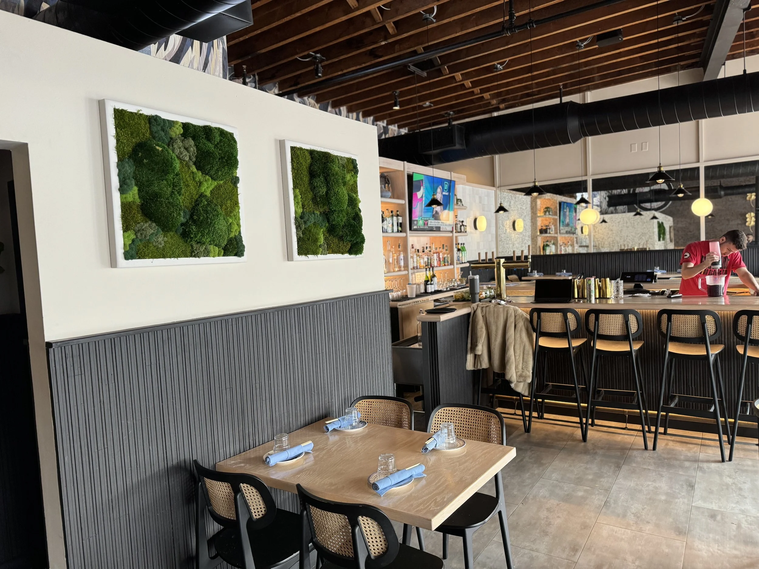 Inside a modern restaurant with a bar area in the background, wooden ceiling beams, wall art with green moss, and a small table set with glasses and napkins.