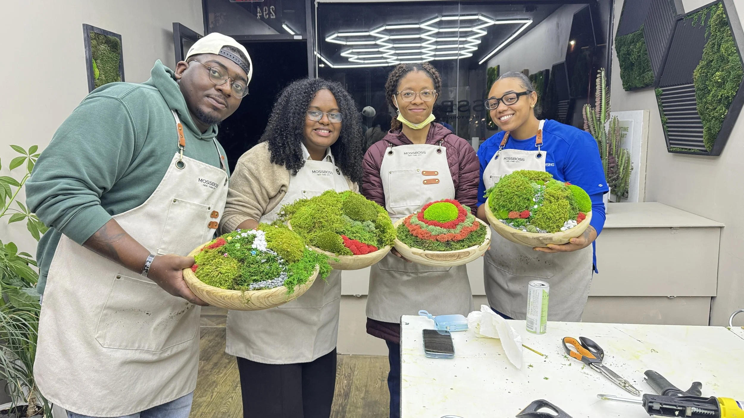 Four people, three women and one man, are standing indoors holding wooden bowls filled with moss and small plants. They are wearing aprons with the Moss Boss logo and are smiling at the camera. The background includes plants, artistic wall decorations, and a table with tools and materials.