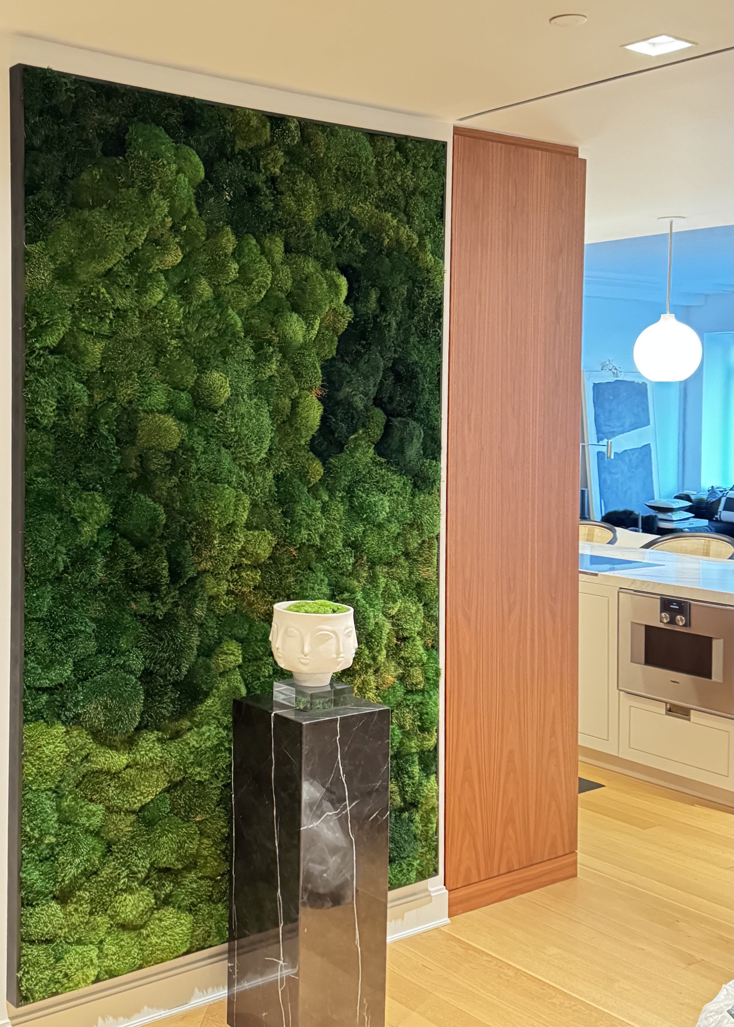 Interior view of a modern room with a green moss wall, a white decorative vase with green moss on a black marble pedestal, wooden wall paneling, and part of a kitchen with light-colored cabinetry and a microwave, near a seating area with blue curtain
