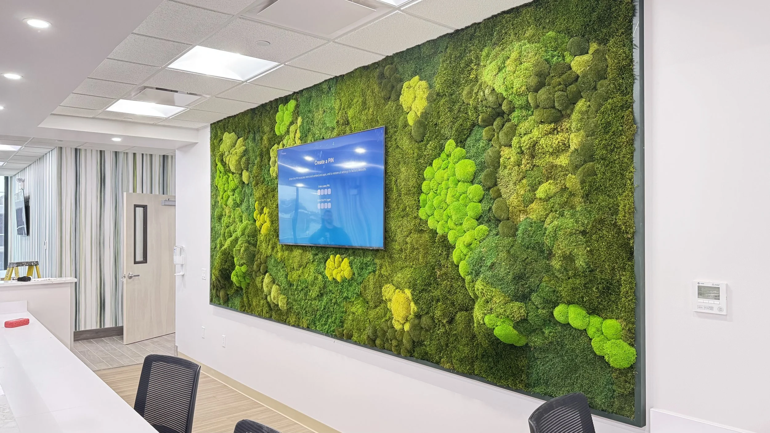 Interior of a modern office conference room with a green moss wall and a mounted digital screen displaying a PIN creation interface.