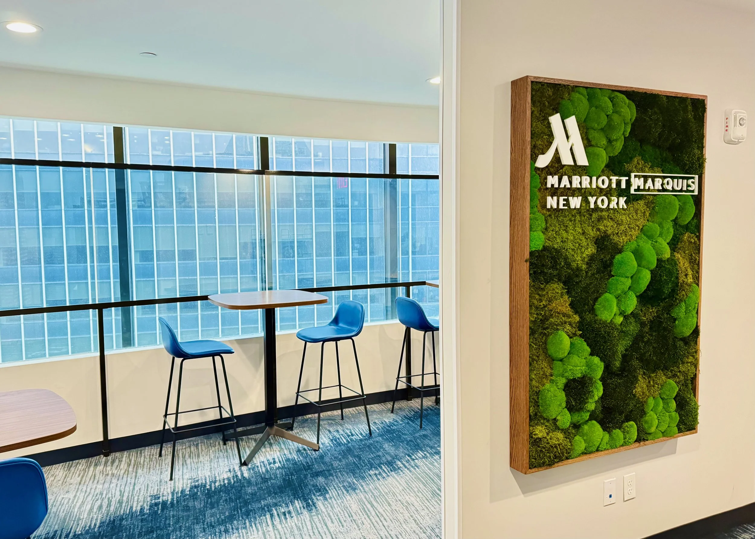 Interior of a Marriott hotel lounge with high tables, blue chairs, large windows, and greenery wall displaying Marriott Marquis New York logo.