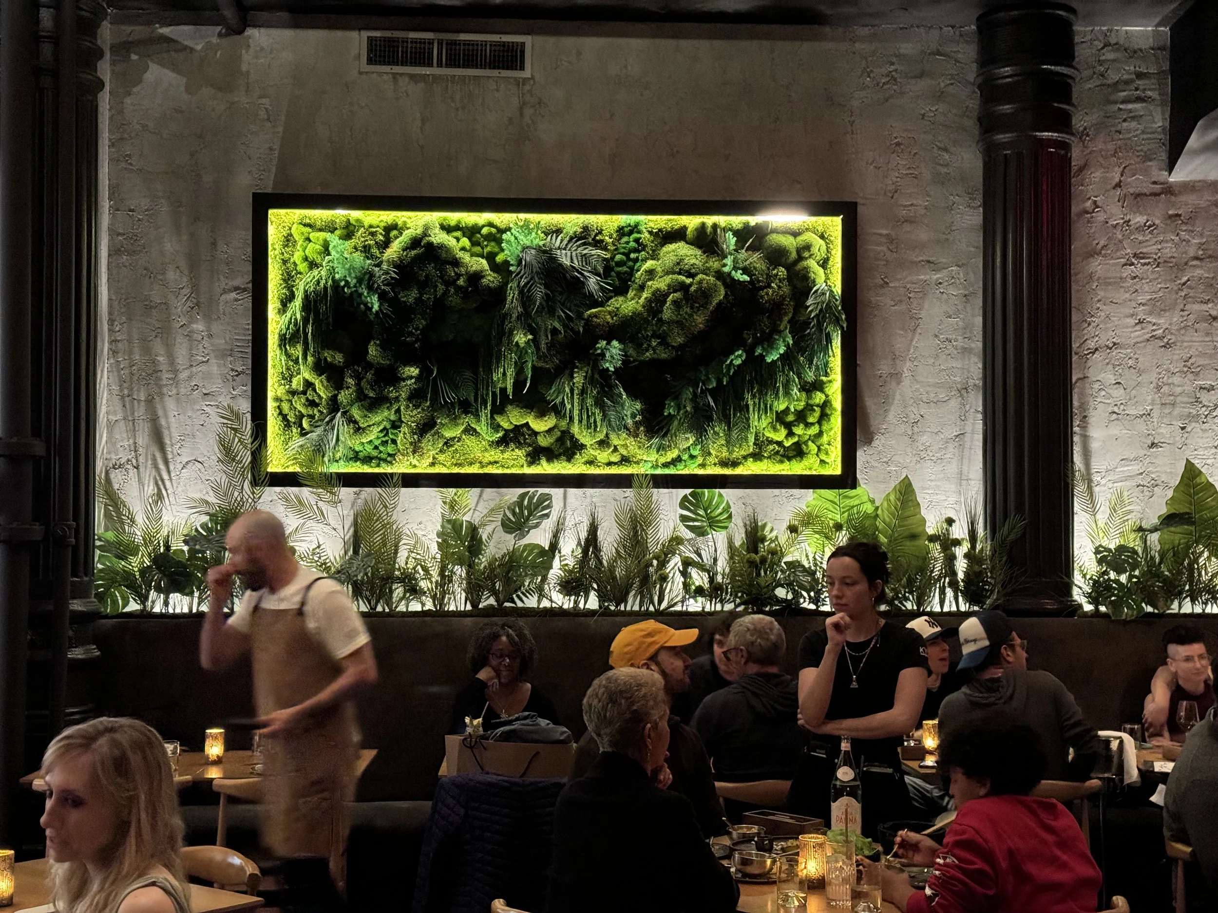 People dining in a dimly lit restaurant with a large green moss and plant wall art