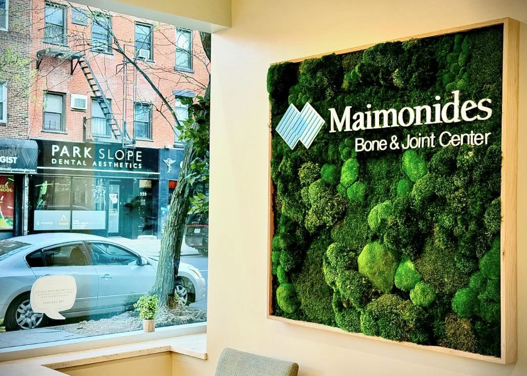 Interior of a medical office with a green moss wall displaying the logo and name of Maimonides Bone & Joint Center. Outside the window, a street scene with parked cars, a tree, and neighboring storefronts, including a dental aesthetics office.