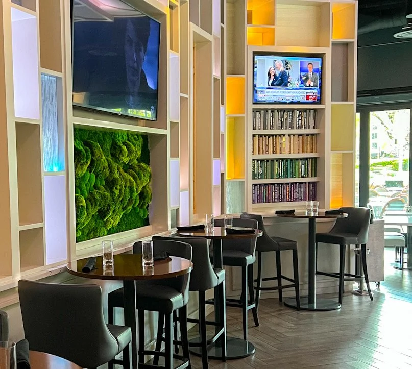 Interior of a modern cafe with high tables and chairs, a wall with bookshelves, flat-screen TVs, and decorative lighting.