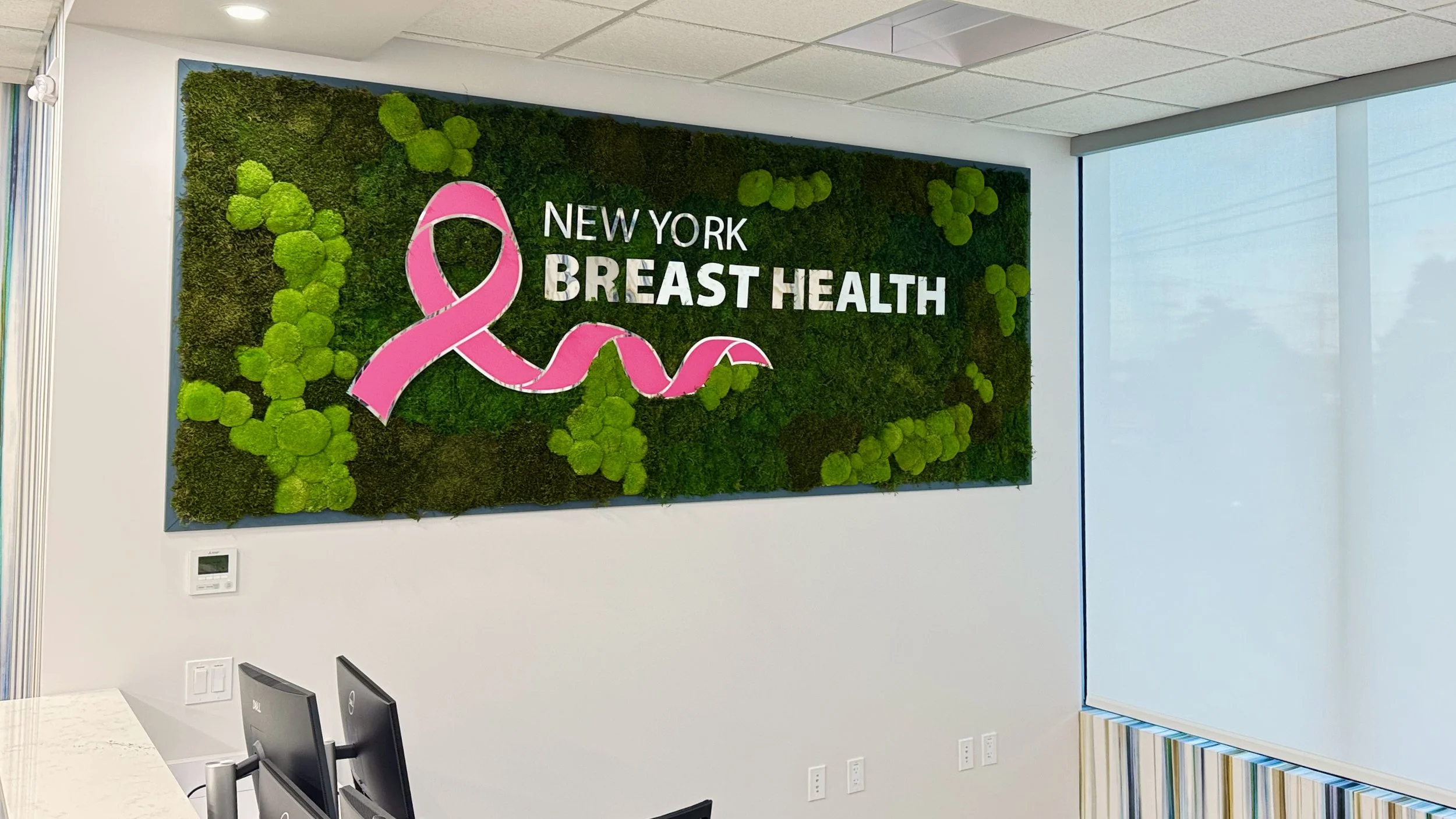 Sign for New York Breast Health with a pink breast cancer ribbon and green foliage background.