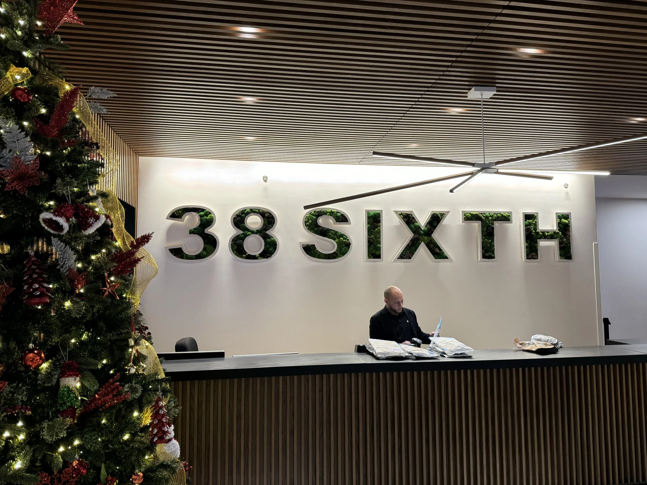 Reception area with a Christmas tree on the left, decorated with ornaments and lights. Behind the counter, a man is working with papers in front of him. The wall behind displays the text '38 SIXTH' with greenery inside the letters, and the ceiling ha