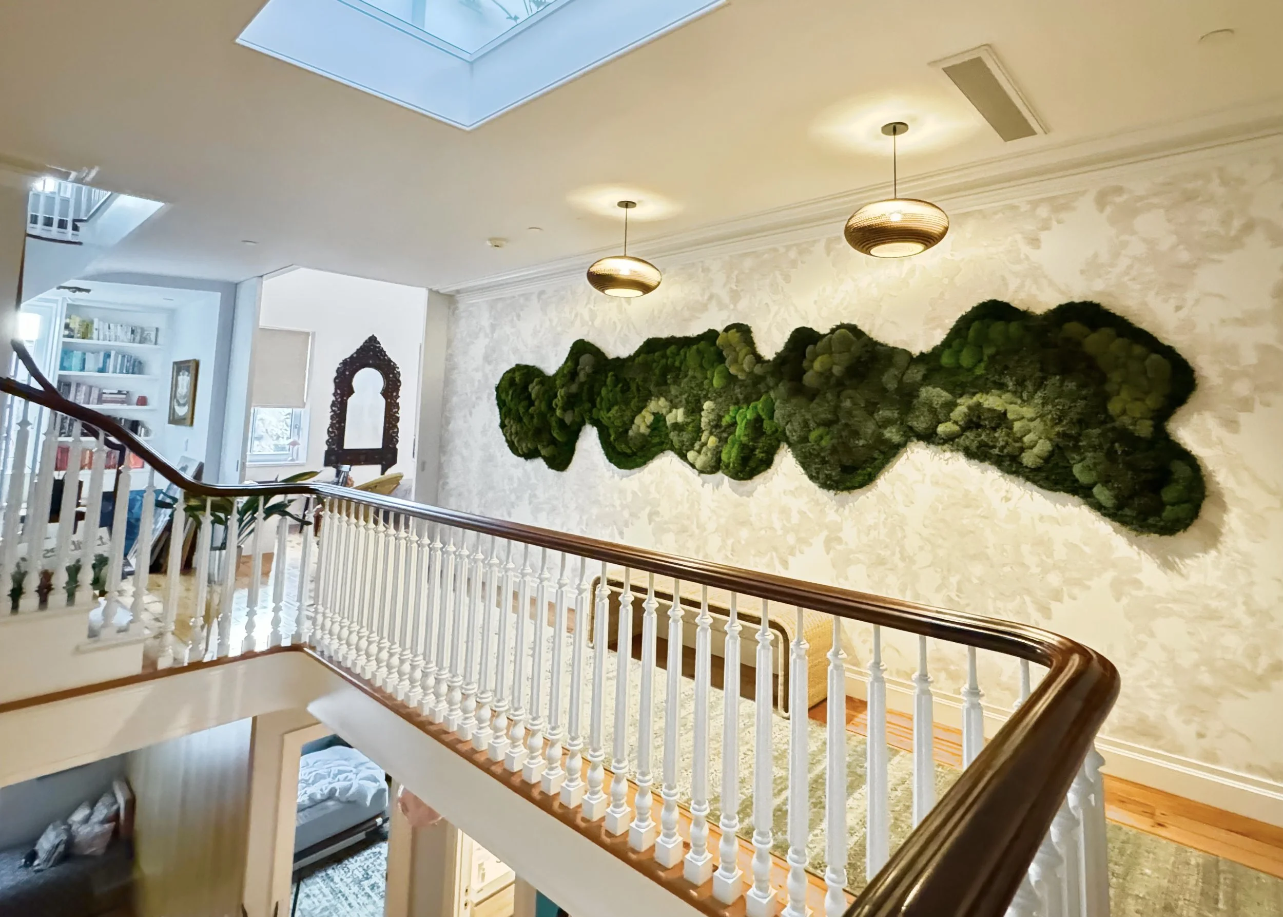 Interior view of a home hallway with a moss wall art installation on the upper wall, two hanging ceiling lights, a wooden handrail, and stairs leading down.
