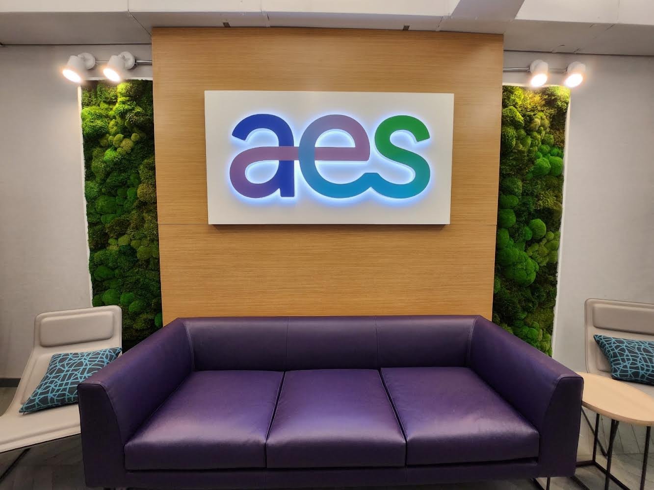 Modern office lobby with a purple sofa, two side chairs with patterned pillows, and a backlit "aes" sign on a wooden accent wall, flanked by green moss walls.