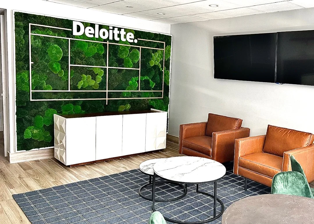 Deloitte office lounge with green moss wall, white cabinet, two tan armchairs, marble coffee tables, black TV screens, and green chairs on wooden floor.